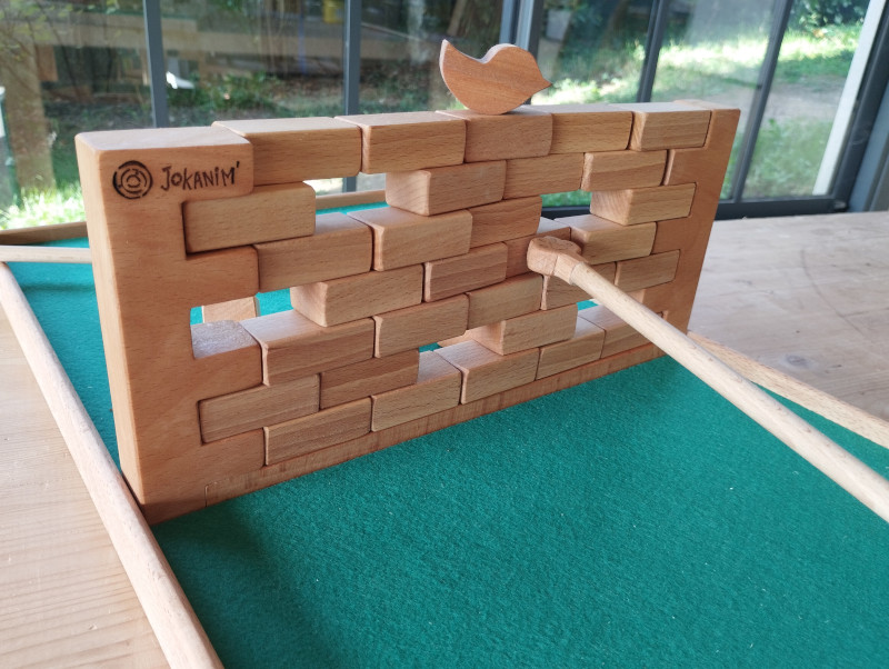 jeu du muret: un oiseau est posé sur un mur de blocs de bois. Une patte de chat, au bout d'un bâton, tente de faire tomber les blocs sans faire tomber l'oiseau.